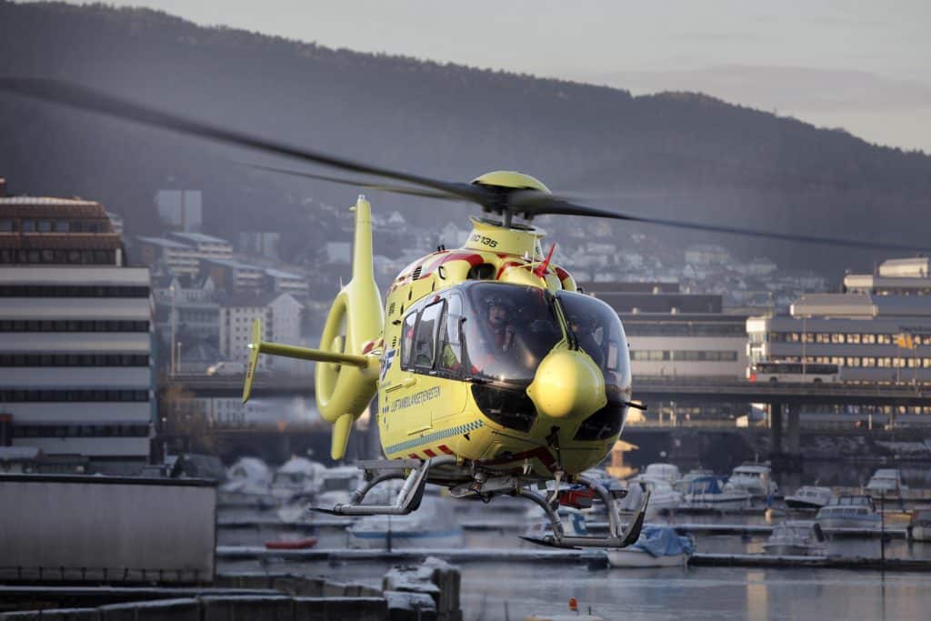 Luftambulansehelikopter i lufta over Bergen by med bygninger og båthavn i bakgrunnen.